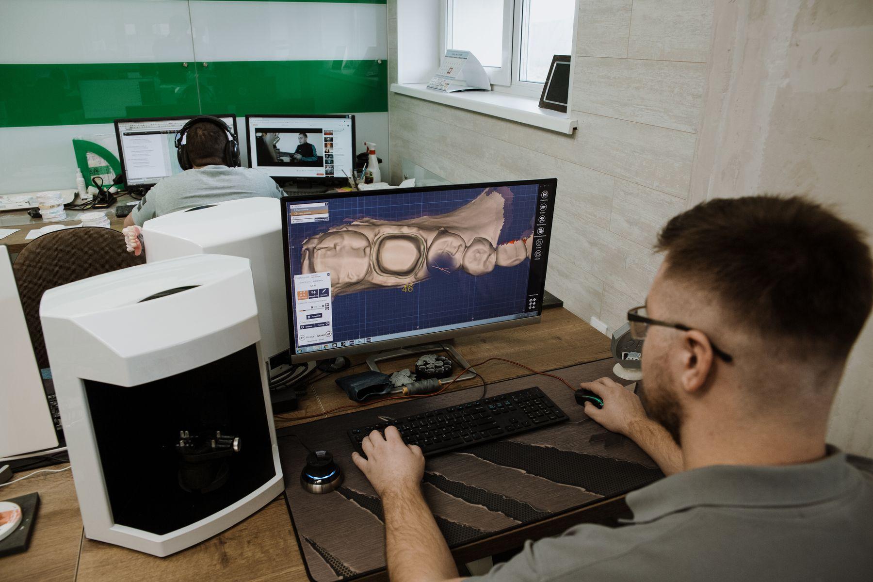 A technician designing an appliance on screen from an intraoral scan, beside a laboratory scanner